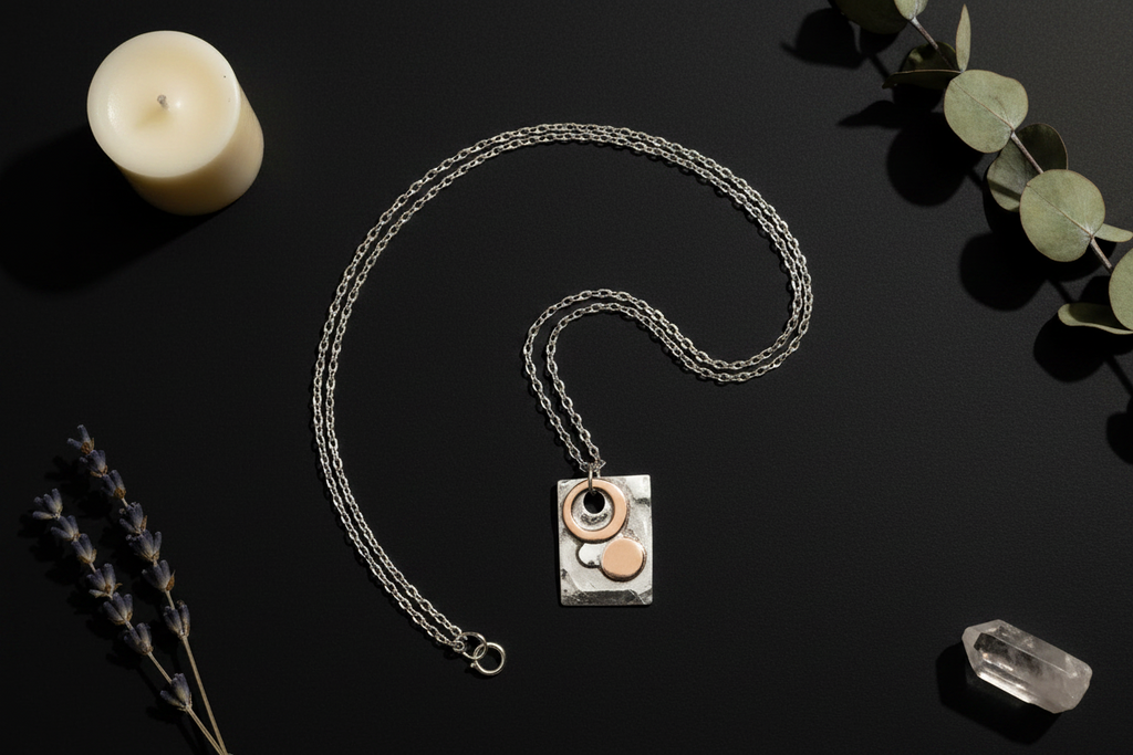 A silver chain necklace with a rectangular silver pendant featuring a hand-drawn style black line drawing of a face, styled on a black background with a candle, eucalyptus leaves, lavender, and a crystal.