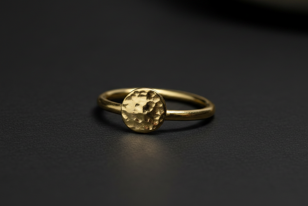 A gold ring with a textured, hammered circular top, resting on a dark, matte surface.