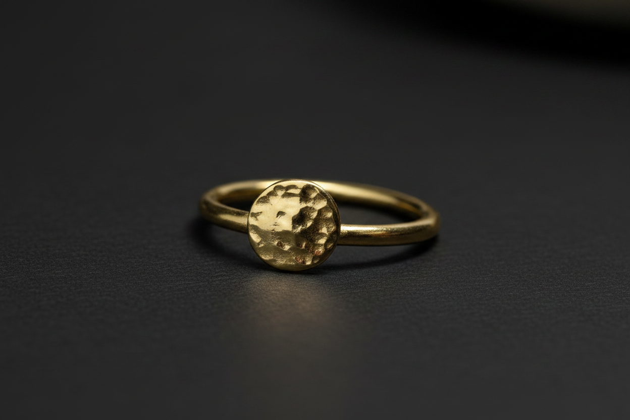 A gold ring with a textured, hammered circular top, resting on a dark, matte surface.
