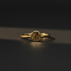 A gold ring with a textured, hammered circular face, resting on a dark, reflective surface under dramatic lighting.