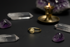 A gold ring with a textured circular face is displayed on a dark surface, surrounded by clear and purple crystals, a brass candle holder with a lit flame, and an amethyst cluster in the background.