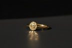 A gold ring with a textured, hammered oval face and a slim band, resting on a dark reflective surface against a black background.