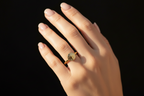 A gold signet ring with a circular, textured face is worn on the ring finger of a hand against a solid black background.