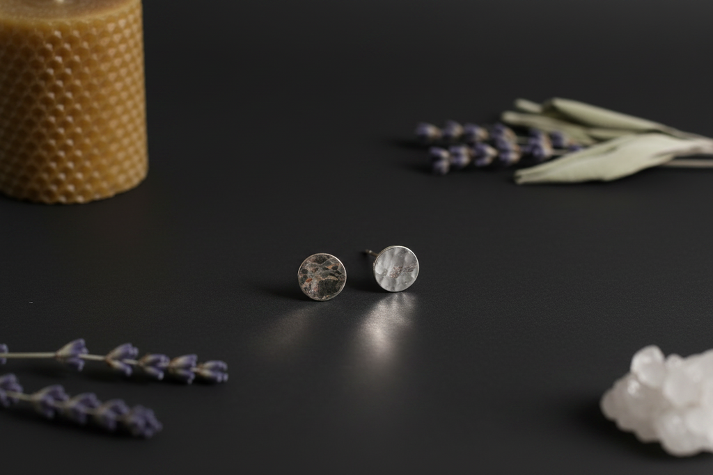 A pair of hammered silver stud earrings displayed on a dark surface, styled with dried lavender sprigs, a textured candle, and white crystals.