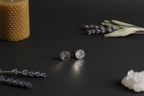 A pair of hammered silver stud earrings displayed on a dark surface, styled with dried lavender sprigs, a textured candle, and white crystals.