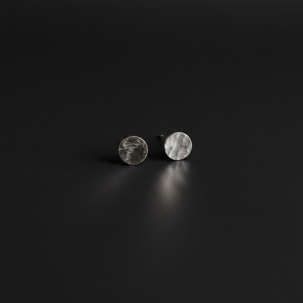 A pair of small round stud earrings with a hammered silver finish, displayed on a dark, reflective surface.