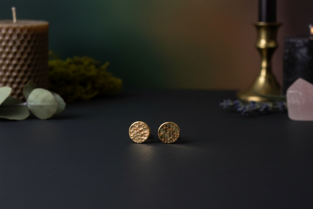 A pair of textured gold stud earrings displayed on a dark surface with a blurred background featuring candles, greenery, and crystals.