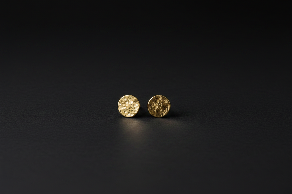 A pair of small round gold stud earrings with a textured hammered finish, displayed on a dark black background.