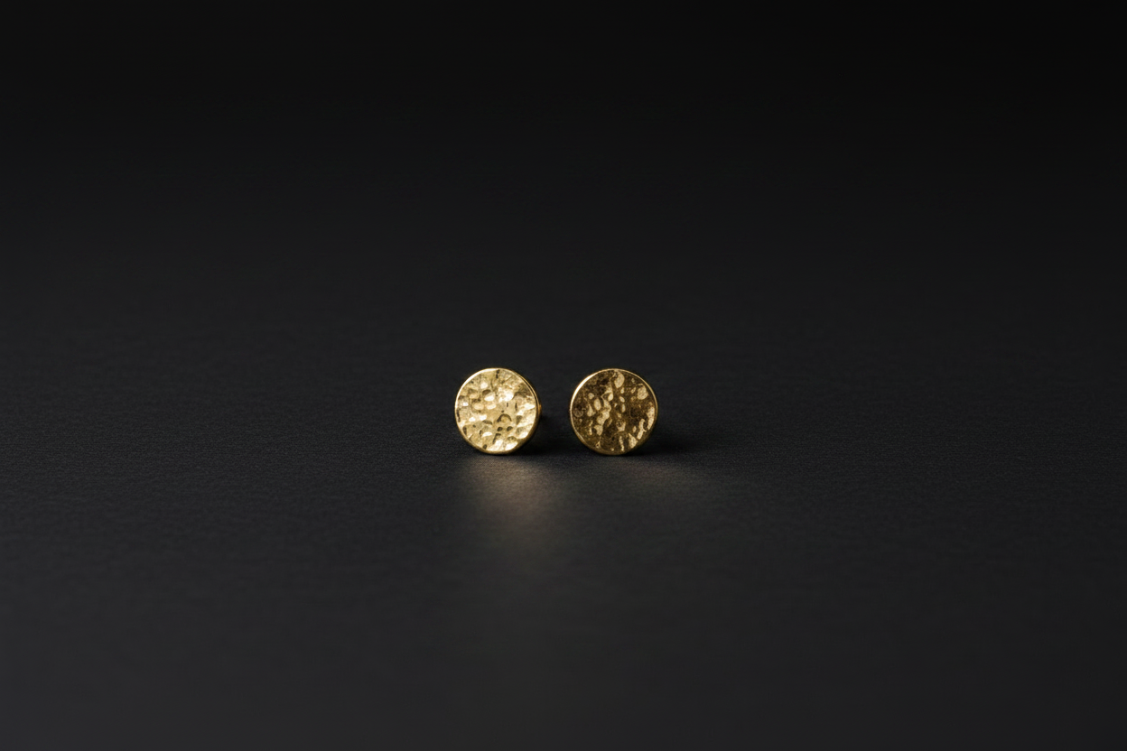 A pair of small round gold stud earrings with a textured hammered finish, displayed on a dark black background.