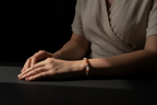 A close-up of a person's hands resting on a dark surface, wearing a delicate beaded bracelet with small peach, white, and dark beads. The person is wearing a light beige linen wrap top against a dark, moody background.