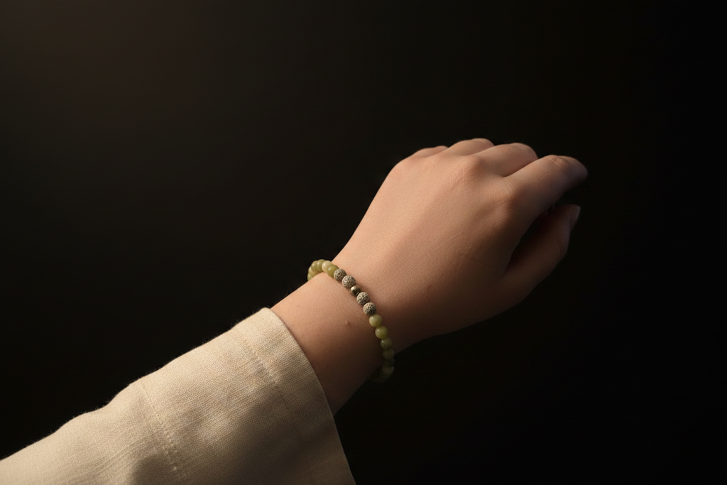 A close-up of a person's wrist wearing a beaded bracelet made of olive green and silver-gray beads, paired with a cream-colored sleeve, set against a dark, solid background.