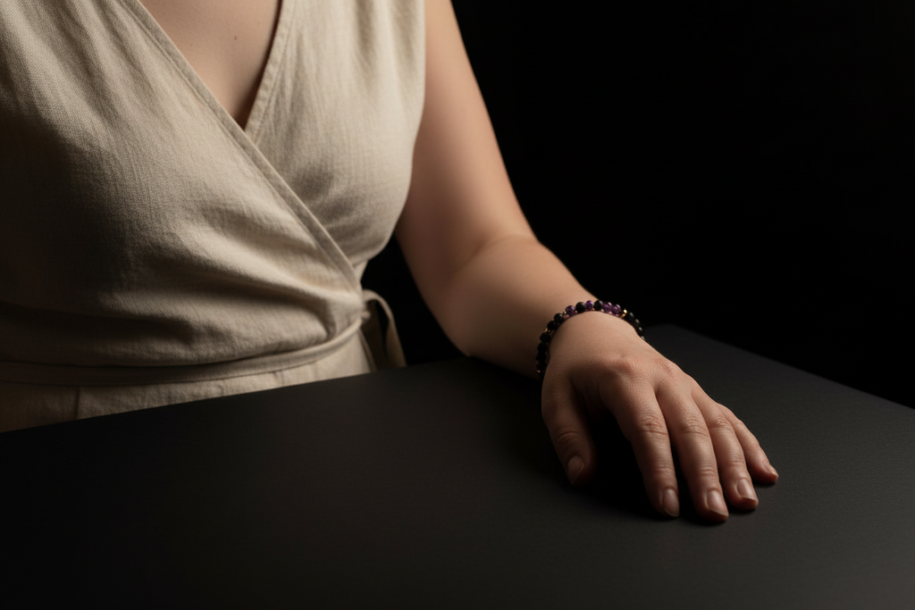 A close-up of a person's hand resting on a dark surface, wearing a dark beaded bracelet. The person is also wearing a light beige wrap-style top, set against a solid black background.