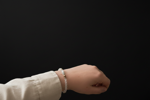 A close-up of a person's wrist wearing a beaded bracelet with white round beads and small metallic gold spacer beads, set against a solid black background.