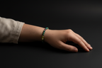 A close-up of a person's hand and wrist wearing a beaded bracelet with green, blue, and yellow beads, set against a solid black background.