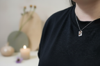 A close-up lifestyle shot of a person wearing a silver-toned necklace with a small square pendant featuring a colorful portrait, paired with a black crewneck sweatshirt. The background is softly blurred with a candle and neutral decor.