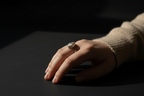 A close-up of a hand wearing a gold ring with a round, textured gray stone, resting on a dark surface under dramatic, low-key lighting.