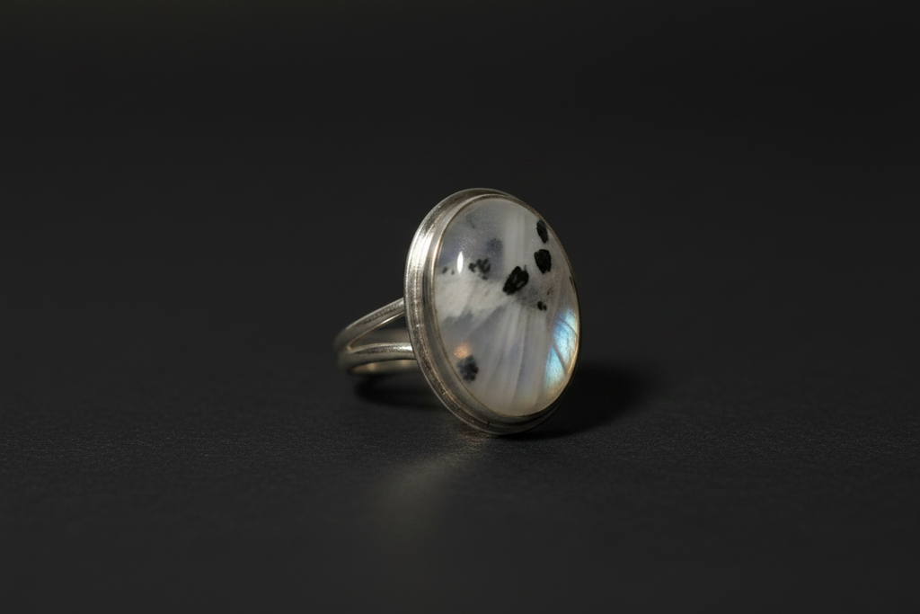 A silver ring featuring an oval cabochon stone with a white and gray marbled pattern and small black speckles, set against a dark background.