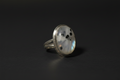 A silver ring featuring an oval cabochon stone with a white and gray marbled pattern and small black speckles, set against a dark background.