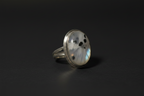 A silver ring featuring an oval cabochon stone with a white and gray marbled pattern and small black speckles, set against a dark background.