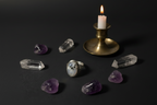 A silver ring with a round white stone featuring black eyes is displayed on a dark surface, surrounded by raw amethyst stones and clear quartz points, with a lit brass candlestick holder in the background.