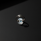 A silver ring with a circular textured disc and a small blue stone in the center, photographed on a dark black background with dramatic lighting.