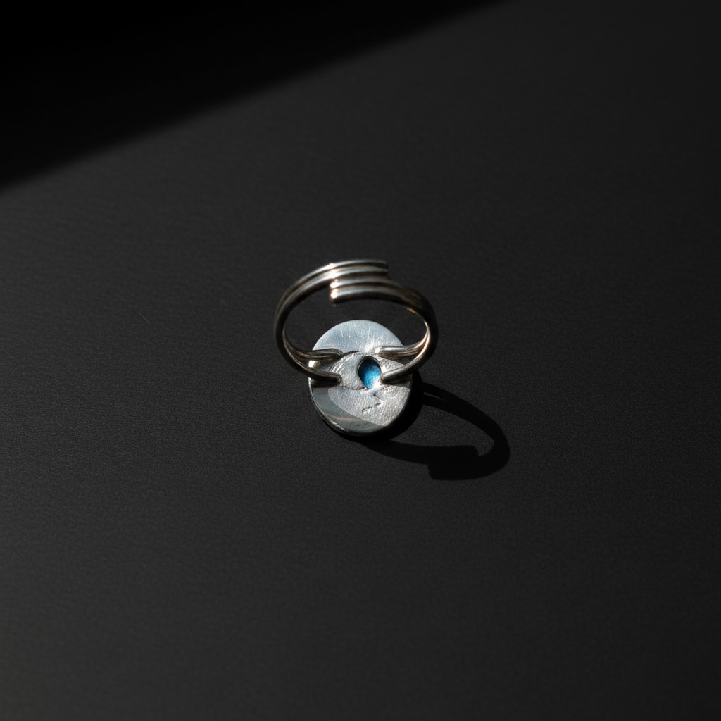 A silver ring with a circular textured disc and a small blue stone in the center, photographed on a dark black background with dramatic lighting.