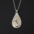 A teardrop-shaped silver pendant necklace featuring a gold inlay and three moon phases set in silver and light blue, suspended from a silver chain against a dark background.