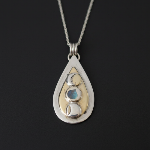 A teardrop-shaped silver pendant necklace featuring a gold inlay and three moon phases set in silver and light blue, suspended from a silver chain against a dark background.