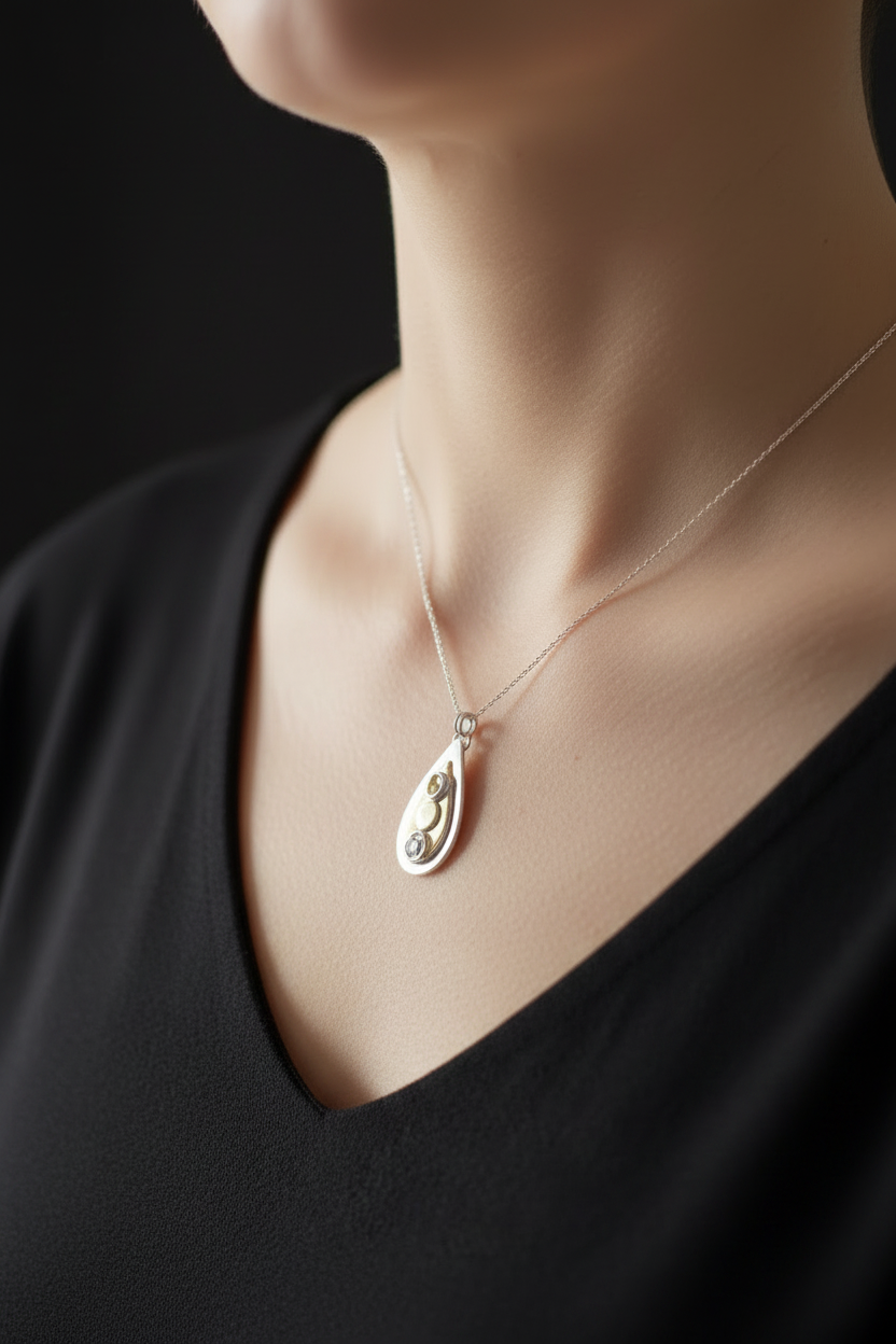 A close-up of a model wearing a delicate silver teardrop pendant necklace with small dark inlaid details, paired with a black V-neck top against a dark background.