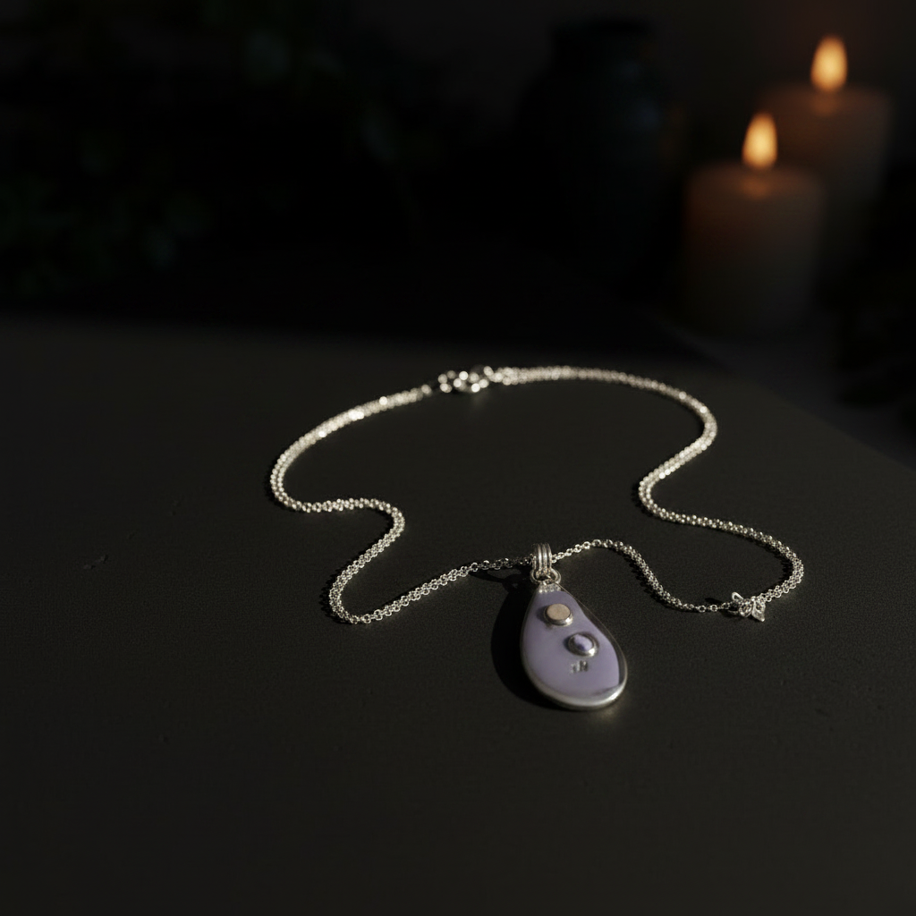 A silver chain necklace with a teardrop-shaped pendant made of pale lavender-gray stone, displayed on a dark surface with softly blurred candles in the background.