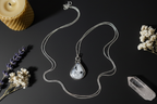 A silver-toned chain necklace with a teardrop pendant featuring a black and white marbled stone, styled on a dark background with dried flowers, a candle, and a crystal.