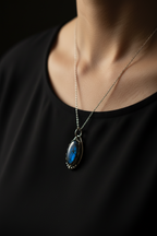 A close-up of a person wearing a silver chain necklace with an oval pendant featuring a vibrant blue iridescent stone, set against a black top and dark background.