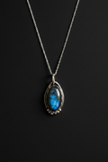 A silver chain necklace with a teardrop-shaped pendant featuring a vibrant blue labradorite stone in a silver bezel setting, displayed against a dark black background.