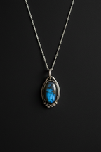 A silver chain necklace with a teardrop-shaped pendant featuring a vibrant blue labradorite stone in a silver bezel setting, displayed against a dark black background.