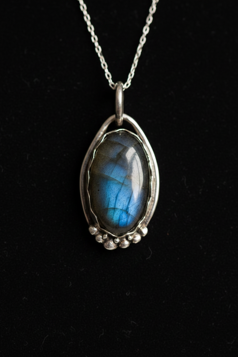 A sterling silver pendant necklace featuring a polished oval labradorite stone with a shimmering blue and green sheen, set against a solid black background.