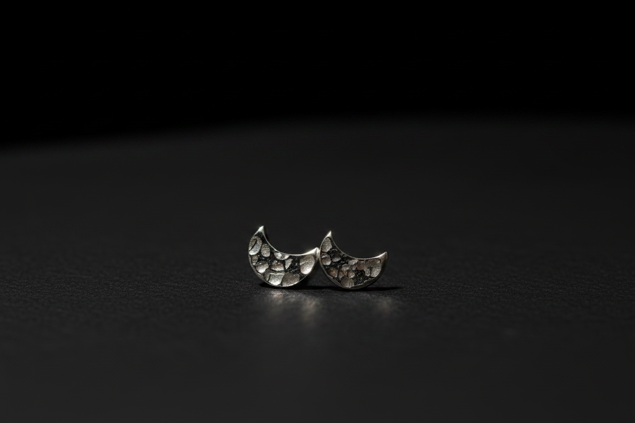 A pair of silver crescent moon stud earrings with a textured, hammered surface, displayed on a dark, reflective surface against a black background.