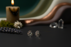 A pair of silver crescent moon stud earrings with textured details, displayed on a dark surface with a lit candle, lavender sprigs, and blurred crystals in the background.