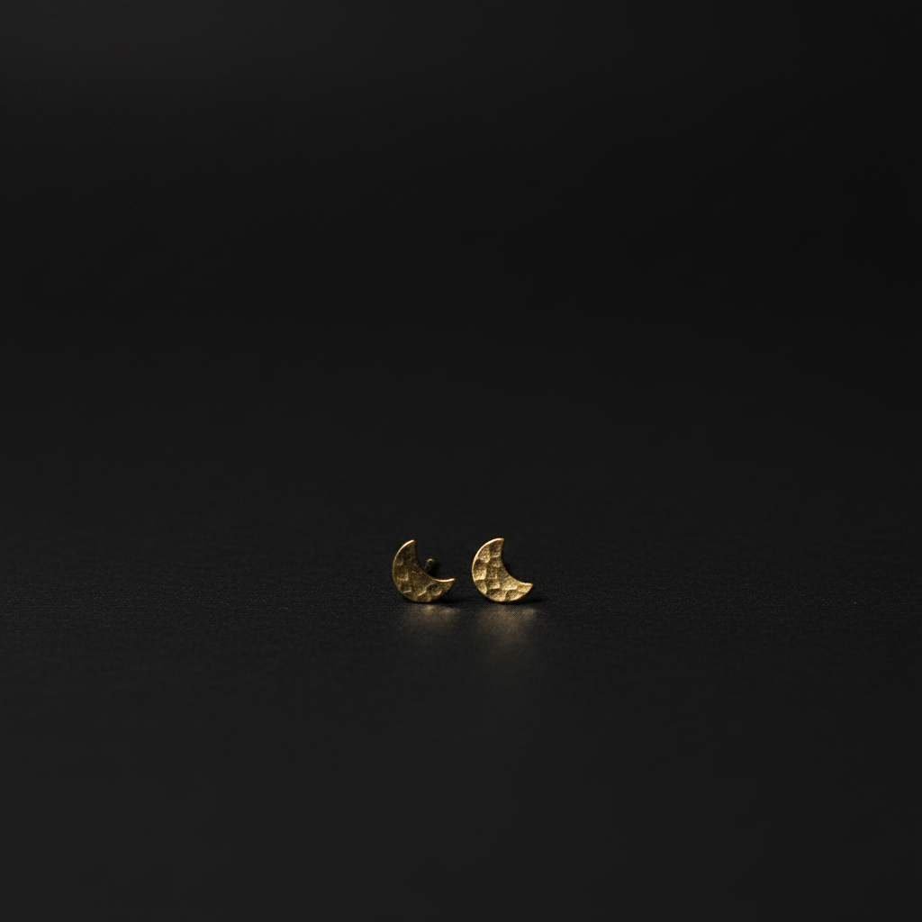 A pair of small crescent moon stud earrings in textured yellow gold, resting on a dark, reflective surface.