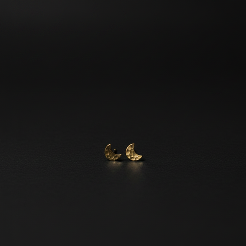 A pair of small crescent moon stud earrings in textured yellow gold, resting on a dark, reflective surface.