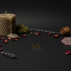 A pair of small gold crescent moon stud earrings displayed on a dark surface, surrounded by decorative props including a textured candle, dried rose petals, lavender sprigs, and a pink crystal.
