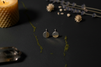 A pair of small round stud earrings with a split silver-tone and gold-tone design, displayed on a dark textured surface with a lit candle, dried flowers, and moss as props.