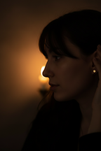 A woman in profile wearing a small gold hoop earring, photographed in very dark lighting with a warm candle glow in the background.