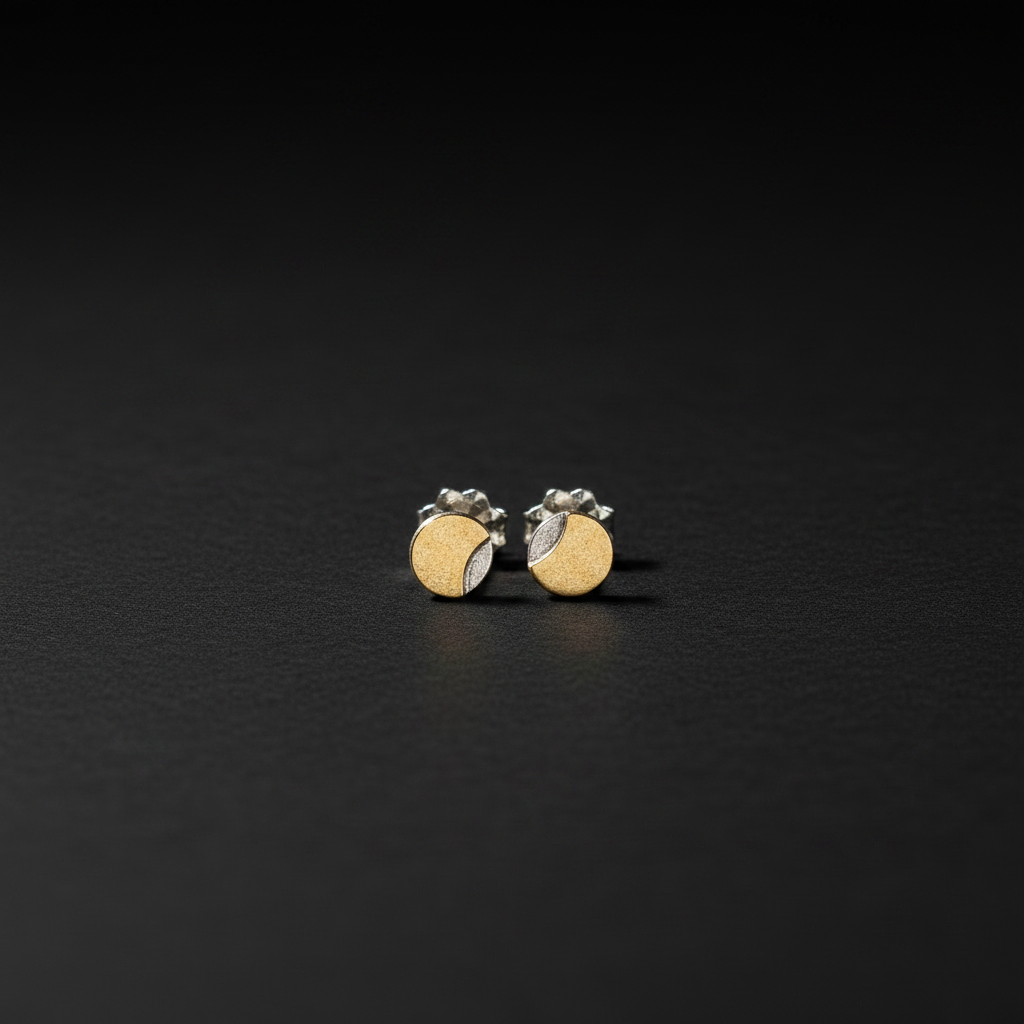A pair of small gold and silver stud earrings with a circular disc design, displayed on a dark textured surface.