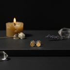 A pair of small gold and silver stud earrings displayed on a dark surface next to a lit candle, crystals, and dried flowers against a black background.
