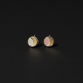 A pair of small round stud earrings on a dark background. Each earring has a silver-toned crescent moon design with one half showing a white textured surface and the other half showing a soft pink textured surface.