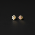 A pair of small round stud earrings on a dark background. Each earring has a silver-toned crescent moon design with one half showing a white textured surface and the other half showing a soft pink textured surface.