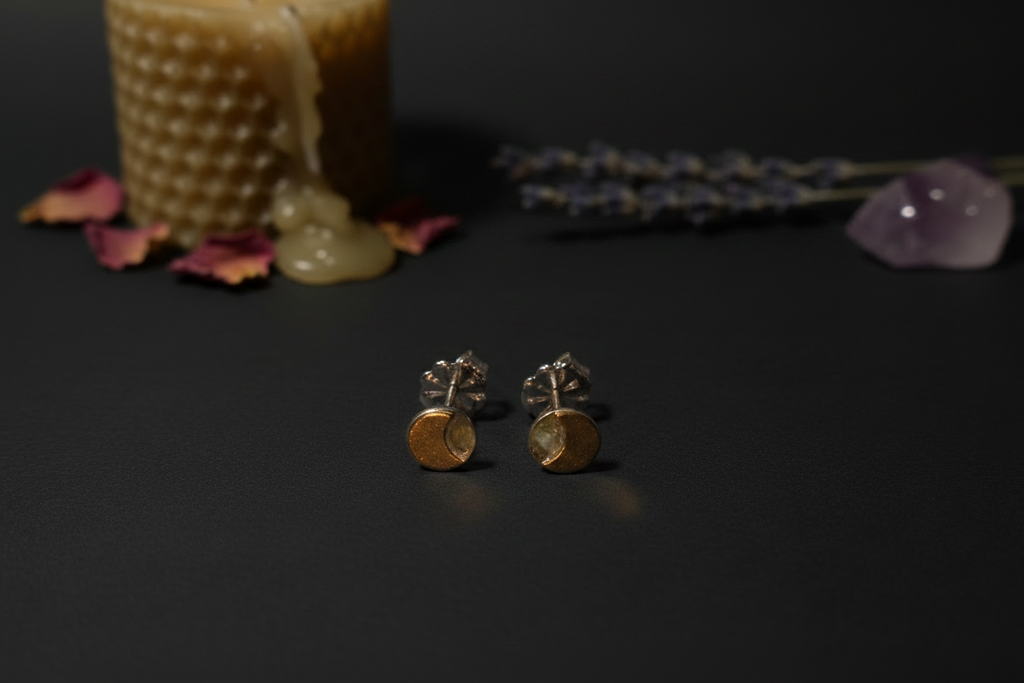 A pair of small gold and silver stud earrings with circular gold disc charms, displayed on a dark surface with a candle, dried lavender, and a crystal in the background.