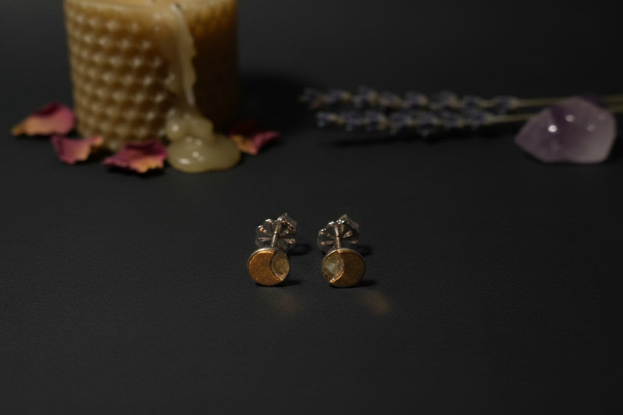 A pair of small gold and silver stud earrings with circular gold disc charms, displayed on a dark surface with a candle, dried lavender, and a crystal in the background.