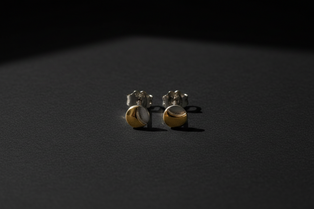 A pair of small gold and silver crescent moon stud earrings resting on a dark, textured surface under dramatic, low-angle lighting.