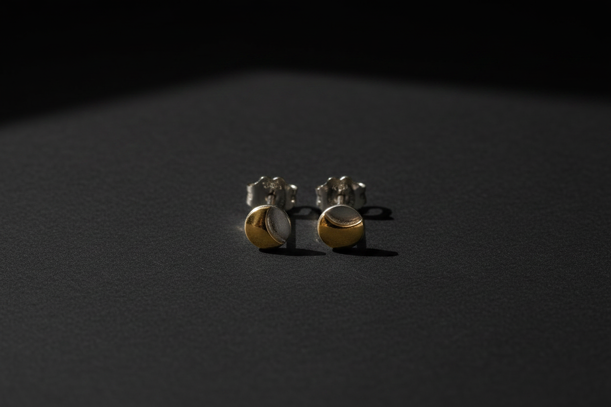 A pair of small gold and silver crescent moon stud earrings resting on a dark, textured surface under dramatic, low-angle lighting.